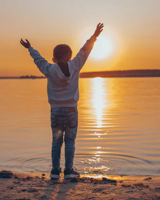 man in white long sleeve shirt and blue denim jeans standing on beach during sunset