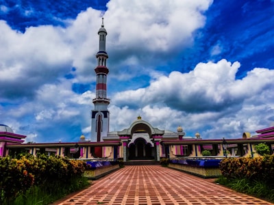 A grand mosque with a tall minaret reaching into a vibrant blue sky filled with puffy white clouds. The intricate architecture features colorful details and symmetrical design. The entrance is framed by lush greenery.