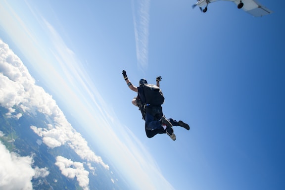 Two people are skydiving, with one person strapped to the back of the other, hovering high above the earth. The sky is a clear blue with some scattered clouds, and the landscape below is slightly visible. An aircraft is seen in the top right corner, suggesting where the jump originated.