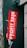 A bold red sticker with white text, placed on a dark green textured metal surface. The text on the sticker reads 'Profit 2020: Money Over Lives', with small text referencing a political statement. Surrounding the sticker are layers of old paper and remnants of previous posters, suggesting an urban environment.