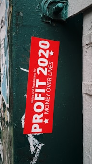 A bold red sticker with white text, placed on a dark green textured metal surface. The text on the sticker reads 'Profit 2020: Money Over Lives', with small text referencing a political statement. Surrounding the sticker are layers of old paper and remnants of previous posters, suggesting an urban environment.