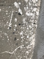 A concrete surface with splatters of white and black paint scattered across it in an abstract pattern. The texture of the concrete is visible, and the paint splatters vary in size, with some appearing as small dots and others as larger, irregular blobs. There are also fine lines of paint running across the surface, adding to the chaotic appearance.