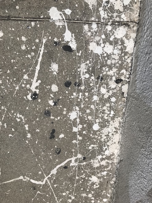 A concrete surface with splatters of white and black paint scattered across it in an abstract pattern. The texture of the concrete is visible, and the paint splatters vary in size, with some appearing as small dots and others as larger, irregular blobs. There are also fine lines of paint running across the surface, adding to the chaotic appearance.