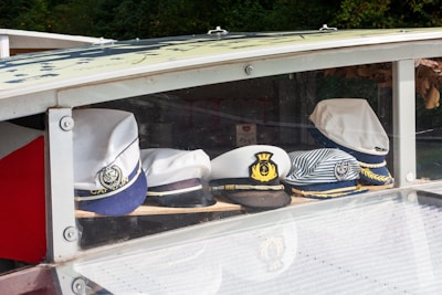 A collection of nautical hats is displayed on a shelf within a glass case. The hats have various designs, including white captain hats with anchor insignias and one with stripes.