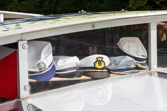 A collection of nautical hats is displayed on a shelf within a glass case. The hats have various designs, including white captain hats with anchor insignias and one with stripes.