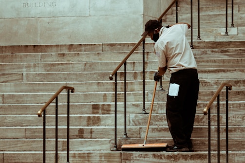 A person wearing a uniform and cap is sweeping a set of stone stairs with a long-handled broom. The background shows part of a building wall with iron railings alongside the stairs.