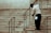 A person wearing a uniform and cap is sweeping a set of stone stairs with a long-handled broom. The background shows part of a building wall with iron railings alongside the stairs.
