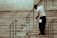 A person wearing a uniform and cap is sweeping a set of stone stairs with a long-handled broom. The background shows part of a building wall with iron railings alongside the stairs.