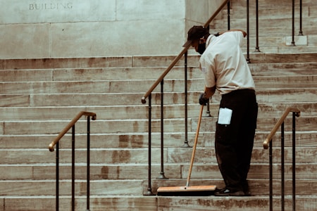 A person wearing a uniform and cap is sweeping a set of stone stairs with a long-handled broom. The background shows part of a building wall with iron railings alongside the stairs.