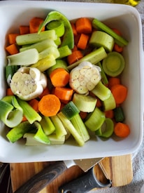 A white ceramic dish filled with peeled and sliced vegetables, including carrots, leeks, celery, and halved garlic bulbs. The vegetables are seasoned with pepper and ready for roasting. The dish is placed on a wooden cutting board, and a black-handled vegetable peeler is visible at the edge.