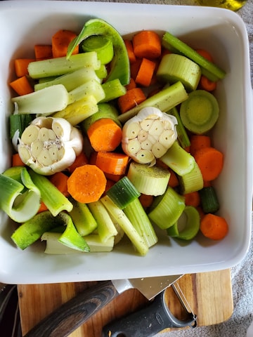 A white ceramic dish filled with peeled and sliced vegetables, including carrots, leeks, celery, and halved garlic bulbs. The vegetables are seasoned with pepper and ready for roasting. The dish is placed on a wooden cutting board, and a black-handled vegetable peeler is visible at the edge.