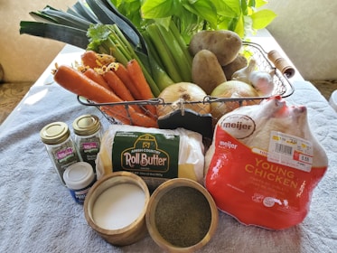 A variety of fresh ingredients is arranged on a kitchen surface. There are carrots, leeks, celery, potatoes, onions, and garlic in a metal basket. In the foreground, there are containers of organic rosemary and thyme, a tub of powder, roll butter, and a packaged young chicken. Nearby, there are small bowls containing salt and a green spice.