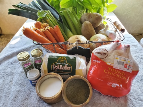 A variety of fresh ingredients is arranged on a kitchen surface. There are carrots, leeks, celery, potatoes, onions, and garlic in a metal basket. In the foreground, there are containers of organic rosemary and thyme, a tub of powder, roll butter, and a packaged young chicken. Nearby, there are small bowls containing salt and a green spice.