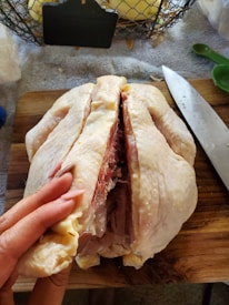 A raw chicken being prepared on a wooden cutting board. It has been partially split open, and a person's hand is holding it open. A large knife is placed beside it. In the background, there are kitchen items including a wire basket with some contents and measuring spoons.
