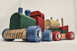 A wooden toy train composed of colorful blocks with sections in green, blue, red, and yellow. The train features small wooden wheels decorated with flower-like patterns and a smiling face on one of the blocks. It is set against a plain background.