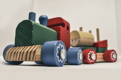 A wooden toy train composed of colorful blocks with sections in green, blue, red, and yellow. The train features small wooden wheels decorated with flower-like patterns and a smiling face on one of the blocks. It is set against a plain background.