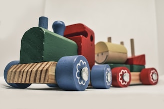 A wooden toy train composed of colorful blocks with sections in green, blue, red, and yellow. The train features small wooden wheels decorated with flower-like patterns and a smiling face on one of the blocks. It is set against a plain background.