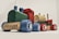 A wooden toy train composed of colorful blocks with sections in green, blue, red, and yellow. The train features small wooden wheels decorated with flower-like patterns and a smiling face on one of the blocks. It is set against a plain background.
