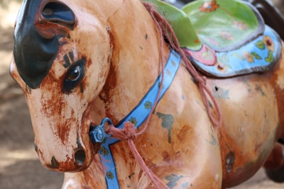 A close-up view of a weathered, vintage rocking horse with chipped paint. The horse features a light brown, wooden finish with visible black markings and scratches. It is adorned with a blue harness decorated with floral patterns and has a green, colorful saddle.