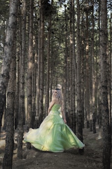 A woman wearing a flowing green dress stands gracefully in a dense forest of tall, slender trees. Her hair is styled in loose curls, and she gazes back over her shoulder, capturing a sense of elegance and mystery. Sunlight filters through the trees, creating dappled light on the forest floor.
