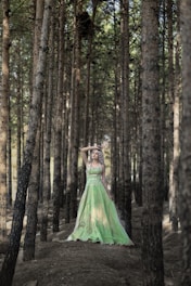 A woman in a flowing green gown stands gracefully in the middle of a dense forest, surrounded by tall, straight trees. Sunlight filters through the leaves, casting dappled patterns on the ground. Her pose is elegant, evoking a sense of tranquility and connection with nature.