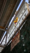 A construction site viewed through metal beams, showing a crane lifting a yellow object against the backdrop of a multi-story building. The perspective is from below, giving an upward view of the structure and machinery.