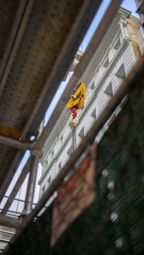 A construction site viewed through metal beams, showing a crane lifting a yellow object against the backdrop of a multi-story building. The perspective is from below, giving an upward view of the structure and machinery.