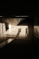 A dimly lit underground passageway features long shadows cast on the tiled floor and walls. The brick-patterned walls are adorned with posters. Light pours in from the entrance, creating stark contrasts between illuminated and shadowed areas. The atmosphere is quiet and somewhat mysterious.