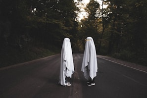 Two figures draped in white sheets stand facing each other on a dark, empty road surrounded by dense trees. The scene has a mysterious and eerie ambiance, with the figures resembling classic representations of ghosts. The surrounding foliage is dense, and the lighting suggests late afternoon or early evening.