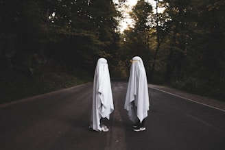 Two figures draped in white sheets stand facing each other on a dark, empty road surrounded by dense trees. The scene has a mysterious and eerie ambiance, with the figures resembling classic representations of ghosts. The surrounding foliage is dense, and the lighting suggests late afternoon or early evening.