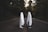 Two figures draped in white sheets stand facing each other on a dark, empty road surrounded by dense trees. The scene has a mysterious and eerie ambiance, with the figures resembling classic representations of ghosts. The surrounding foliage is dense, and the lighting suggests late afternoon or early evening.