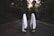 Two figures draped in white sheets stand facing each other on a dark, empty road surrounded by dense trees. The scene has a mysterious and eerie ambiance, with the figures resembling classic representations of ghosts. The surrounding foliage is dense, and the lighting suggests late afternoon or early evening.