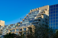 An apartment building with multiple floors, each having balconies with glass railings. The structure has a tiered design, with each floor slightly recessed, creating a cascading effect. There is another modern building with a glass facade adjacent to it. Trees with green foliage are visible in the foreground, and the sky is clear and blue.