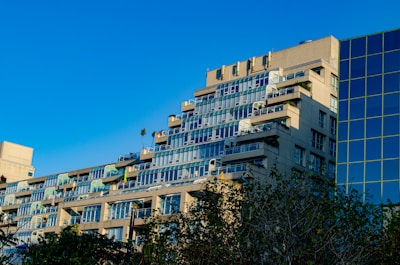 An apartment building with multiple floors, each having balconies with glass railings. The structure has a tiered design, with each floor slightly recessed, creating a cascading effect. There is another modern building with a glass facade adjacent to it. Trees with green foliage are visible in the foreground, and the sky is clear and blue.