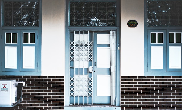 A building entrance with a central door featuring both a glass panel and a metallic gate. The door and surrounding window frames are painted blue, while the walls are white and the lower part of the façade is covered with dark brick tiles. There are two windows on each side of the door, one of them partly displaying a Mitsubishi air conditioning unit. Above the door, a house number plaque displays the number 387.