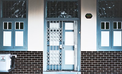 A building entrance with a central door featuring both a glass panel and a metallic gate. The door and surrounding window frames are painted blue, while the walls are white and the lower part of the fa&ccedil;ade is covered with dark brick tiles. There are two windows on each side of the door, one of them partly displaying a Mitsubishi air conditioning unit. Above the door, a house number plaque displays the number 387.