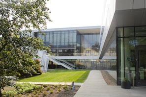 A modern building with large glass windows and a sleek design is situated amidst greenery. The structure features an outdoor staircase and is surrounded by well-maintained grass and trees. The setting appears to be tranquil and well-organized, ideal for an academic or corporate environment.