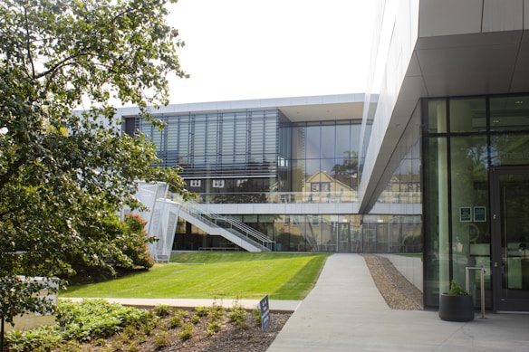 A modern building with large glass windows and a sleek design is situated amidst greenery. The structure features an outdoor staircase and is surrounded by well-maintained grass and trees. The setting appears to be tranquil and well-organized, ideal for an academic or corporate environment.