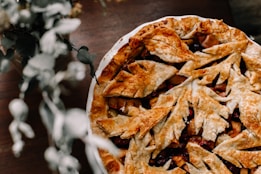 A freshly baked pie with a golden-brown crust intricately designed with leaf patterns, sitting on a wooden table. Parts of the filling, composed of sliced fruits, are visible through the lattice of the crust. A blurred bouquet of flowers is present in the foreground, adding a touch of greenery.