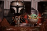 A stylized figurine of a character in futuristic armor stands beside a small, green, large-eared creature in a floating crib. The scene is set on a rocky base with a 'Star Wars' logo, and there is a blurred background of additional merchandise.