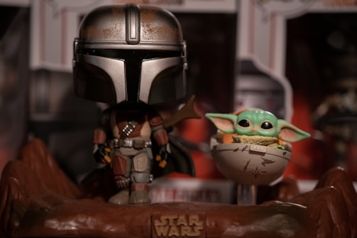 A stylized figurine of a character in futuristic armor stands beside a small, green, large-eared creature in a floating crib. The scene is set on a rocky base with a 'Star Wars' logo, and there is a blurred background of additional merchandise.