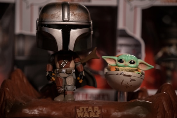A stylized figurine of a character in futuristic armor stands beside a small, green, large-eared creature in a floating crib. The scene is set on a rocky base with a 'Star Wars' logo, and there is a blurred background of additional merchandise.