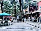 A lively outdoor dining area lined with tall palm trees, featuring several people sitting at tables beneath large umbrellas. The setting is an open plaza with a paved walkway, and a cyclist is passing through the center. The surrounding buildings have a vibrant aesthetic with businesses visible in the background.