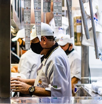 A man wearing a white uniform and black mask works in what appears to be a busy kitchen or meat packing area. Several printed order slips hang above him. Another person in a similar outfit is visible in the background.
