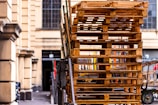 A large stack of wooden pallets is piled up on the side of a street next to a building with arched windows. The scene appears to be set in an urban environment with some bicycles visible in the background.