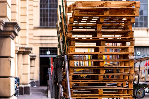 A large stack of wooden pallets is piled up on the side of a street next to a building with arched windows. The scene appears to be set in an urban environment with some bicycles visible in the background.