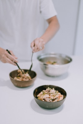 A person is preparing or serving food, using utensils to mix ingredients in ceramic bowls on a white surface. The image captures a close-up view with a focus on the contents of the bowls, which appear to contain a mixture of vegetables or salad.