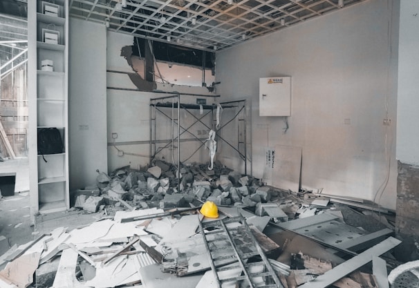 A construction or renovation site filled with debris and rubble. There is a ladder on the ground and scattered building materials, including a hard hat prominently visible in the center. The walls show signs of damage, and a new ceiling structure is partially constructed. Shelves on the left side hold items and a backpack. A maintenance box with a caution sign is attached to the wall.