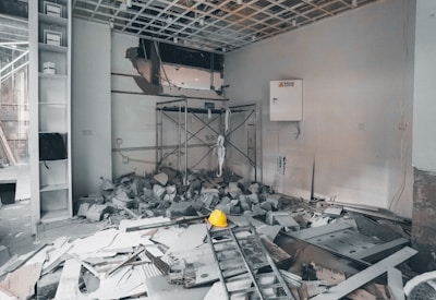 A construction or renovation site filled with debris and rubble. There is a ladder on the ground and scattered building materials, including a hard hat prominently visible in the center. The walls show signs of damage, and a new ceiling structure is partially constructed. Shelves on the left side hold items and a backpack. A maintenance box with a caution sign is attached to the wall.