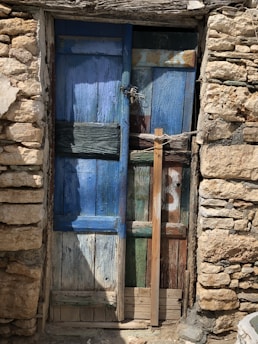 A rustic, weathered wooden door with a patchwork of blue, green, and brown panels, flanked by roughly hewn stone walls. A wooden plank is used as reinforcement across the door.